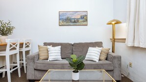 Una sala de estar con un sofá gris, cojines blancos, una mesita con una planta, un cuadro en la pared y una lámpara de pie.
