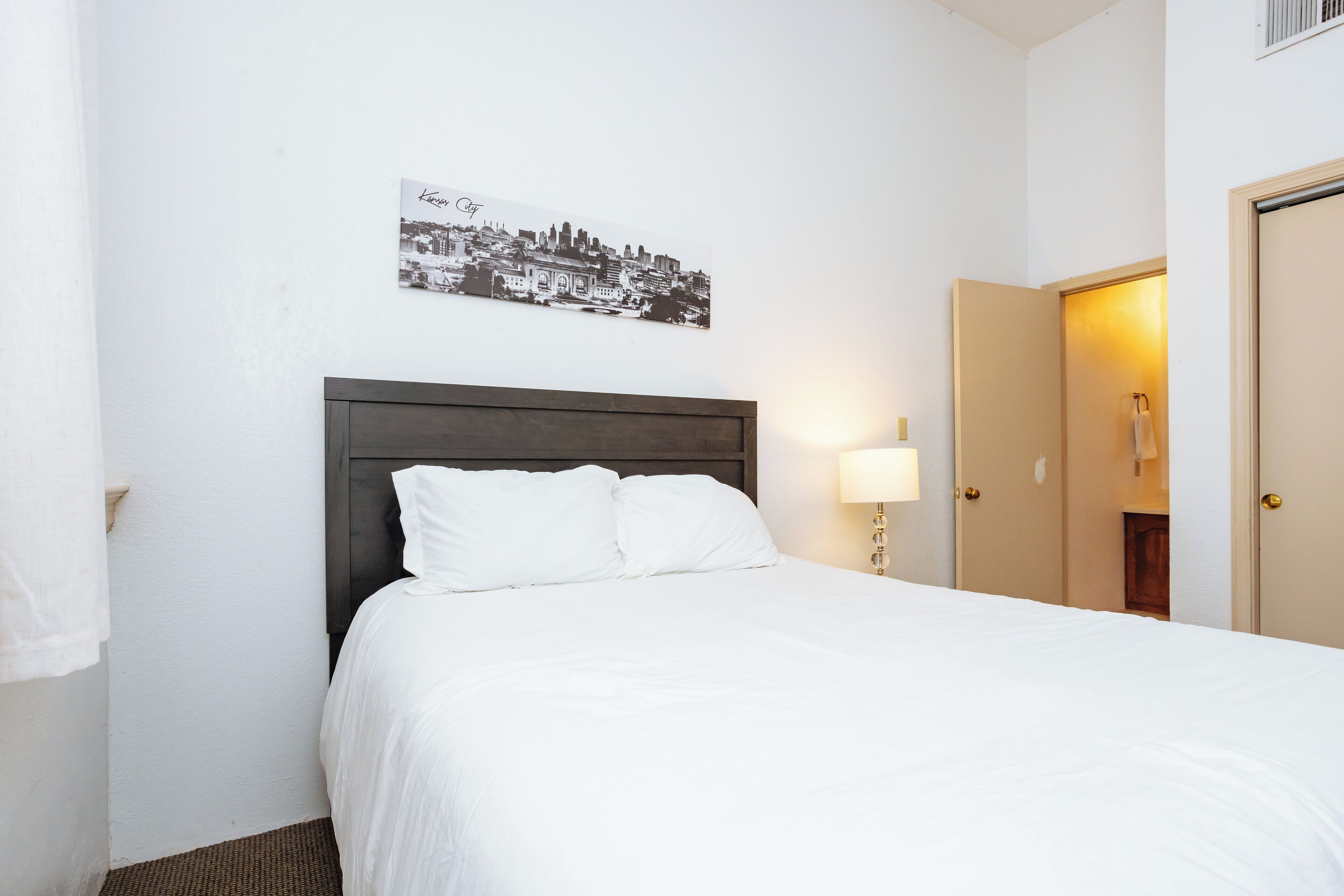 Una cama bien hecha con sábanas blancas, cabecera de madera oscura y un cuadro colgado en la pared con una vista urbana.