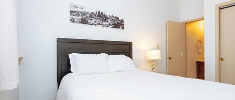 Una cama bien hecha con sábanas blancas, cabecera de madera oscura y un cuadro colgado en la pared con una vista urbana.