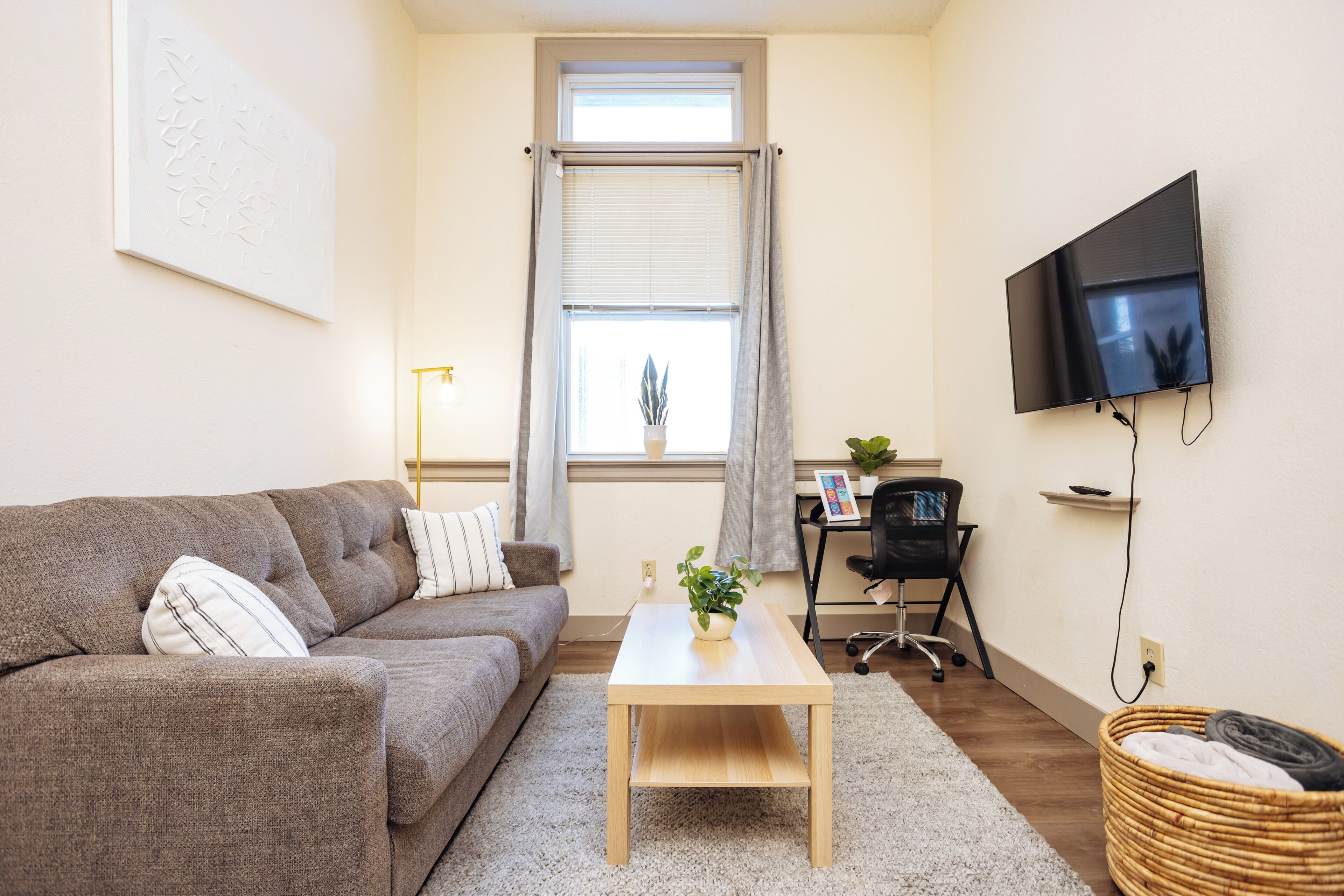 Een woonkamer met een bank, een salontafel, een aan de muur bevestigde televisie, een raam met gordijnen en een klein bureau met een stoel.