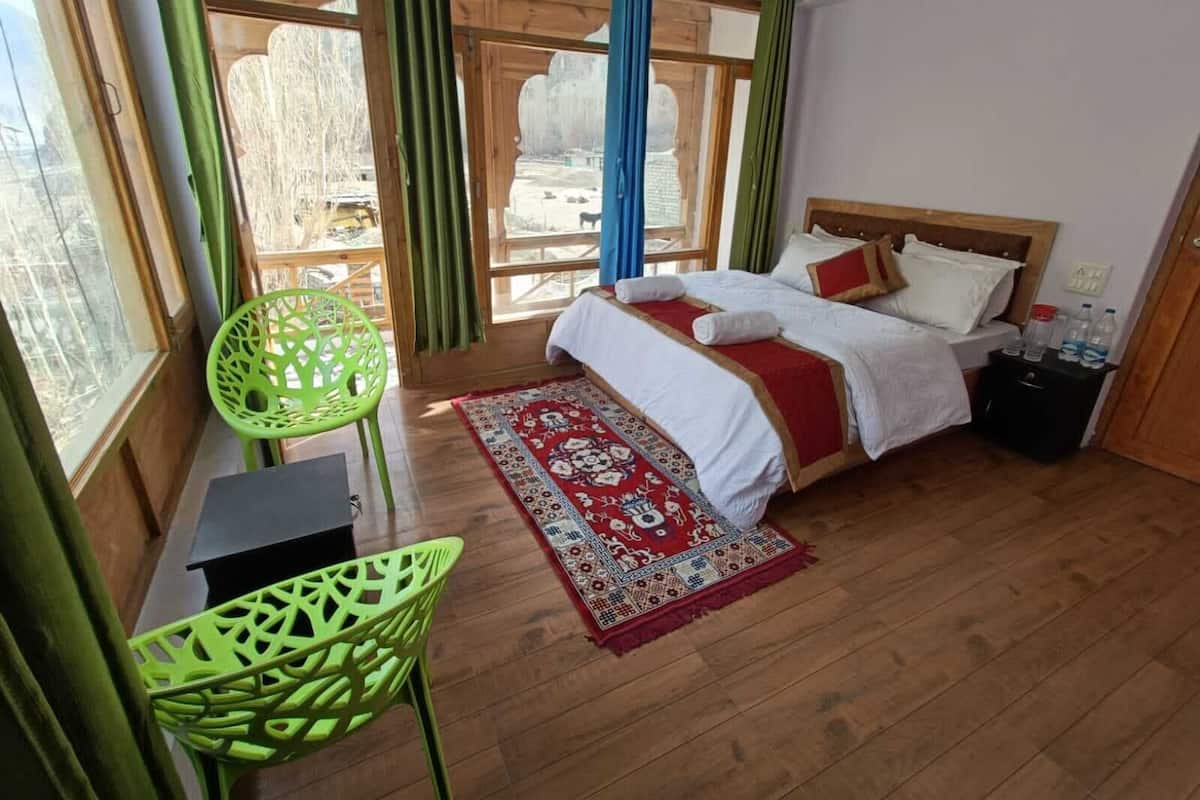 A bedroom with a wooden bed, a red and white patterned rug, a green chair, and a view of a snowy landscape through large windows.