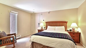 A bedroom with a large bed, two bedside tables with lamps, a wooden nightstand, a chair, and a door. - The Birch Ridge: Mission Room #4 - Queen Suite in Killington, Vermont (Killington)