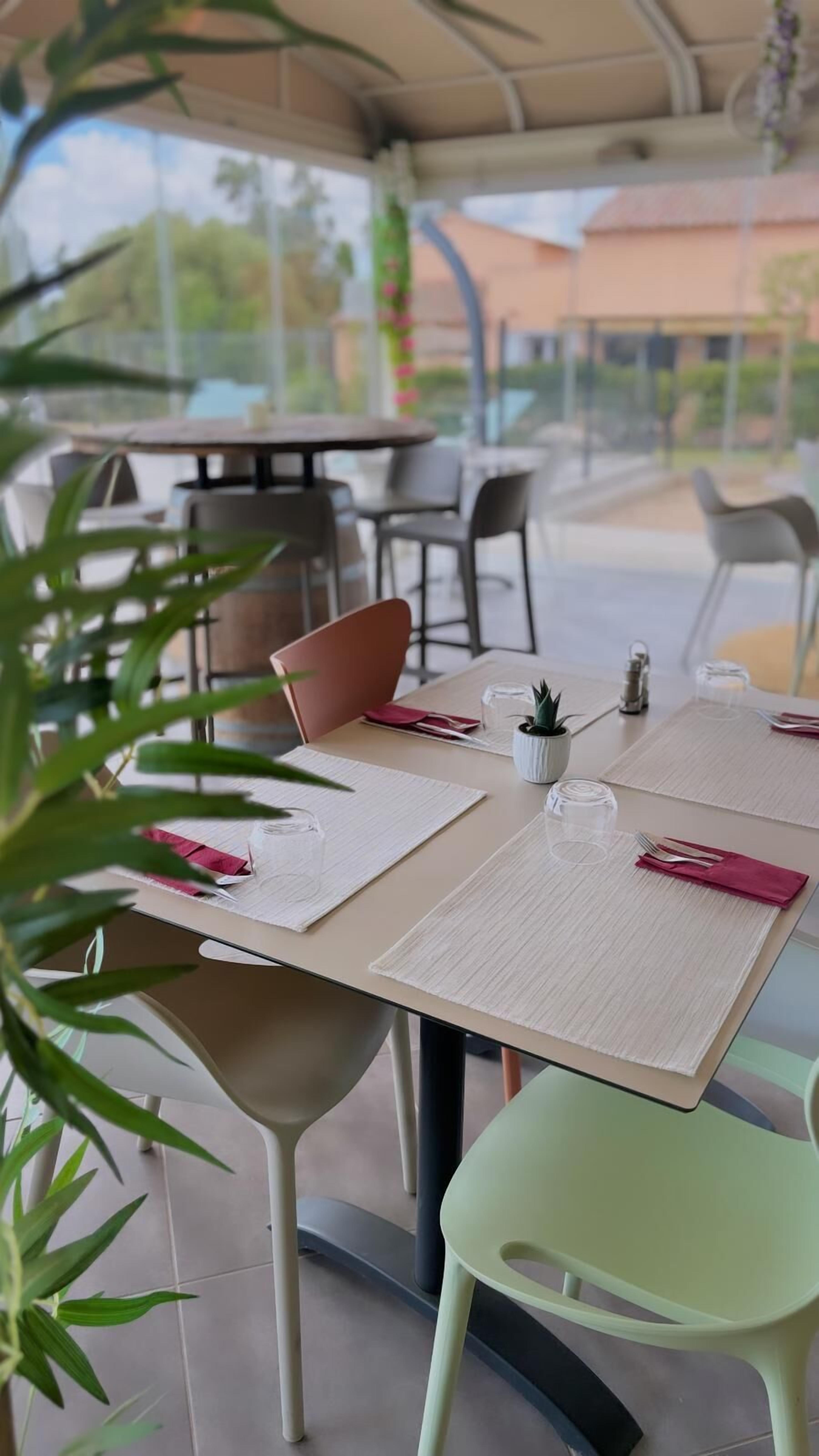 An outdoor dining area with tables set for two, featuring a mix of modern and classic chairs, a small potted plant, and a clear view of the surrounding greenery and buildings.