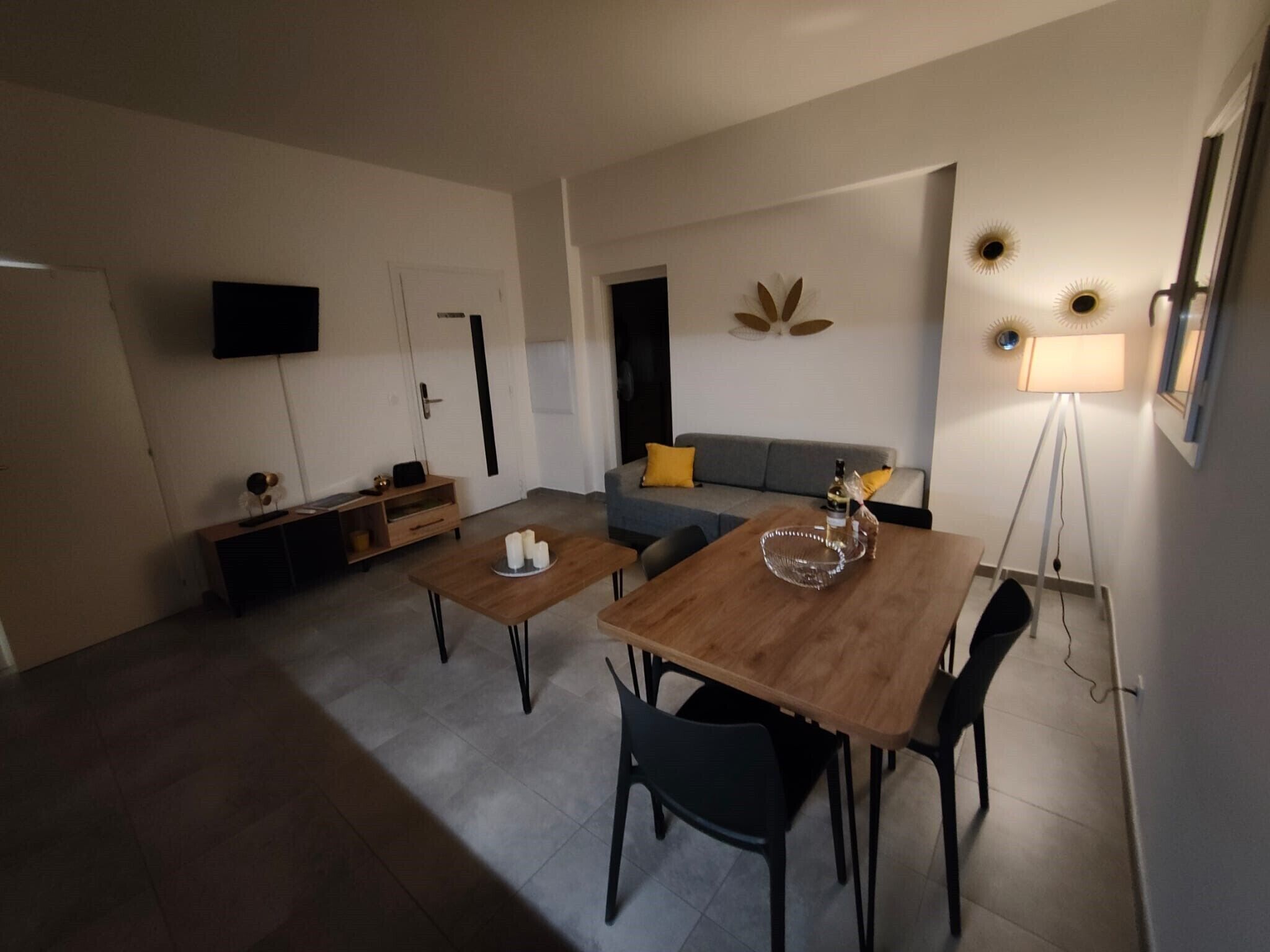 A modern living room with a dining area, grey sofa, and a flat-screen TV mounted on the wall.