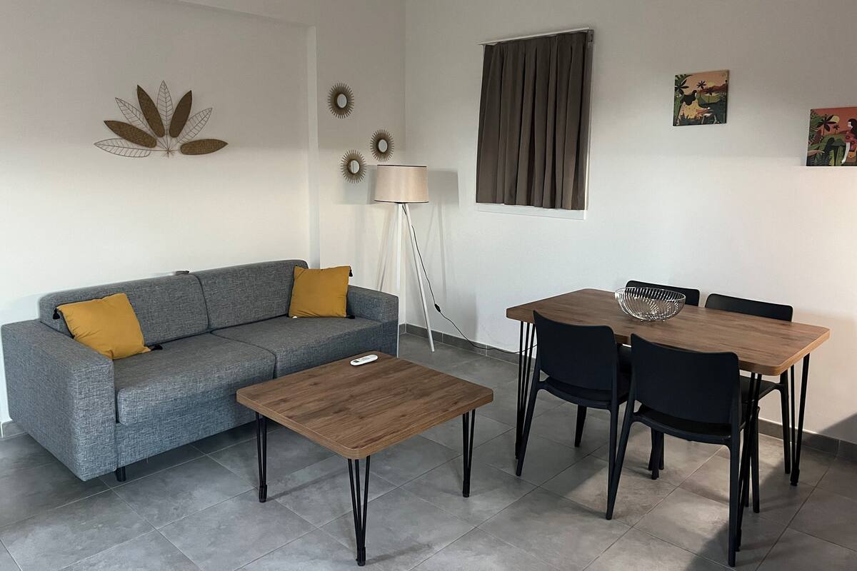 A modern living room with a grey sofa, a wooden coffee table, a dining table with four chairs, a floor lamp, and wall art.