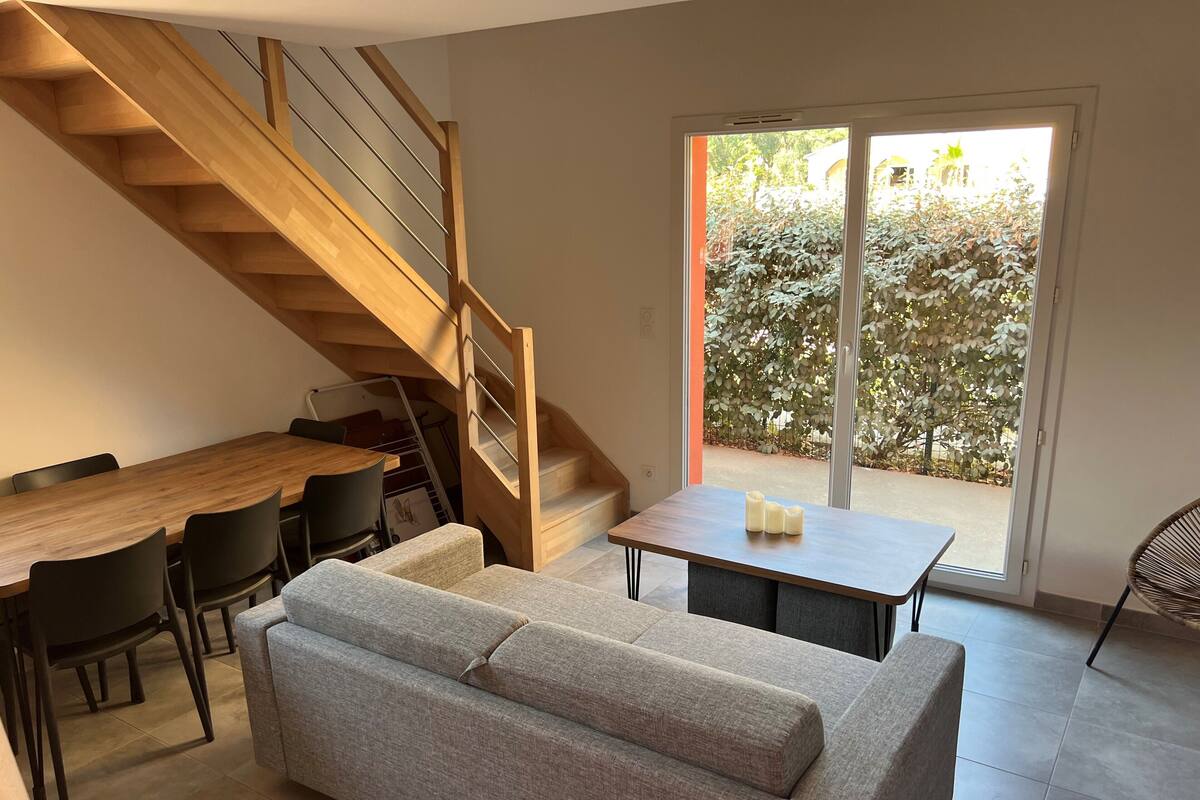 A modern living space with a wooden staircase, a dining area with a wooden table and chairs, a grey sofa, and a sliding glass door leading to an outdoor area.