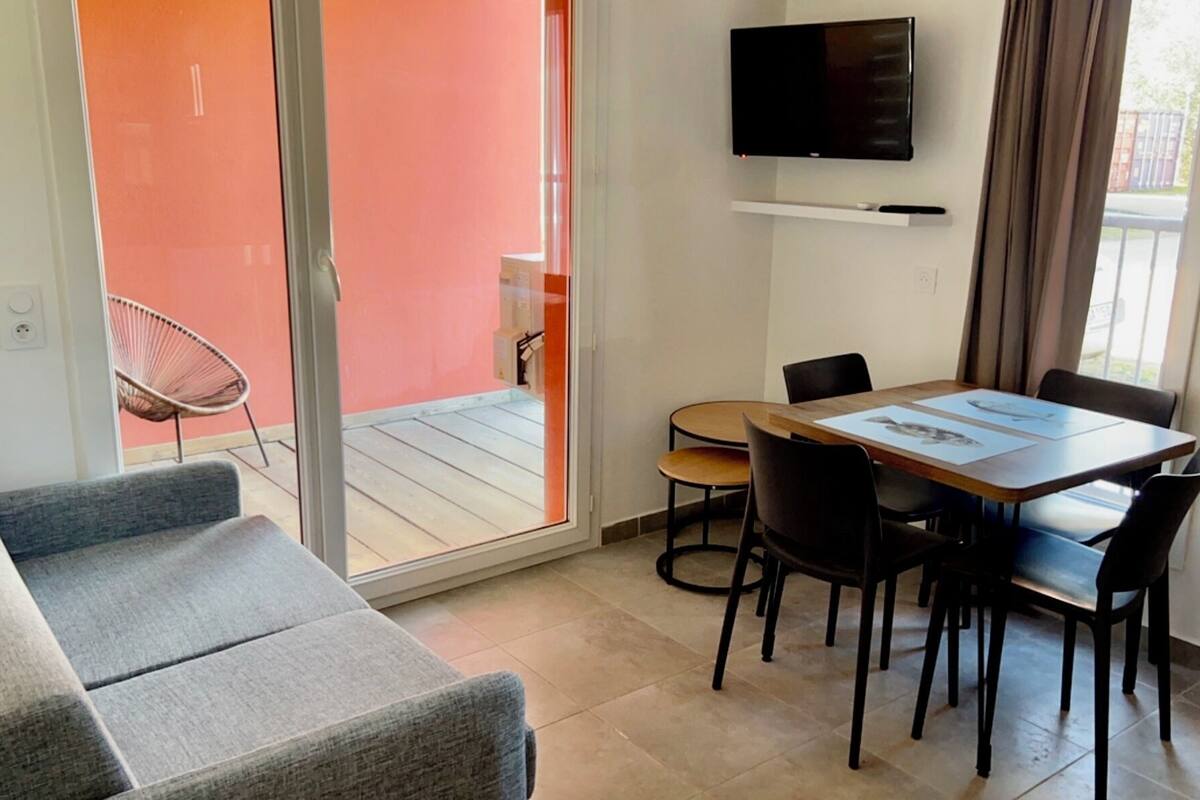 A modern living room with a grey sofa, a dining table with chairs, a flat-screen TV mounted on the wall, and a sliding glass door leading to an outdoor area.