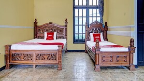 Two wooden beds with intricate carvings, a window with blue shutters, and a door in a room with yellow walls. - SPOT ON 91798 Hotel Grand Mutiara (Cikampek)