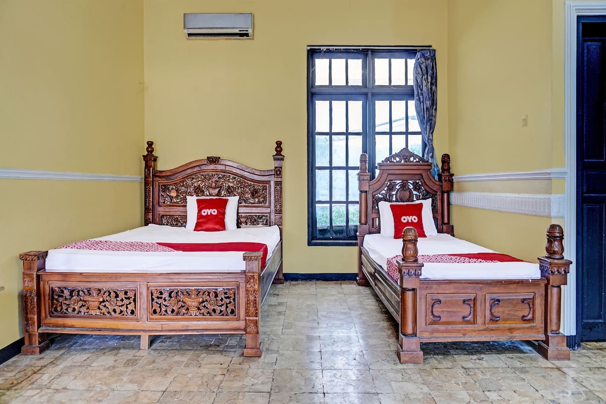 Two wooden beds with intricate carvings, a window with blue shutters, and a door in a room with yellow walls.
