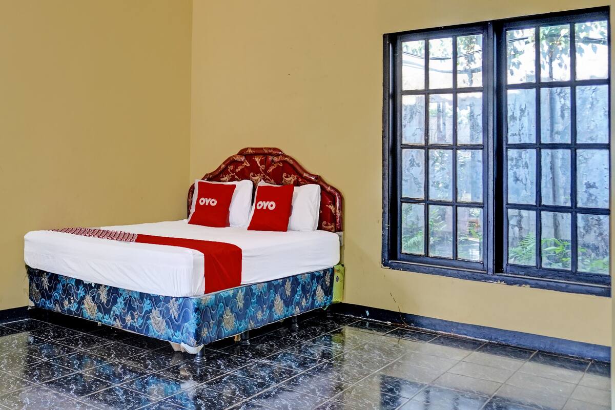 A neatly made bed with a patterned bedspread, two red pillows with the word "OYO" on them, and a window with a view of greenery.