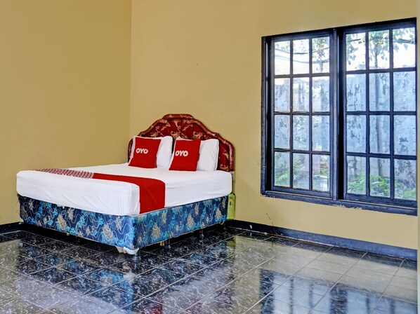 A neatly made bed with a patterned bedspread, two red pillows with the word "OYO" on them, and a window with a view of greenery.