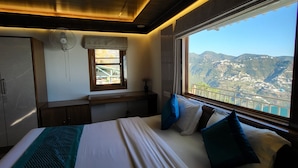 A bedroom with a large bed, a window with a mountain view, a desk, and a wardrobe. - Saiva Hill Resort (Dehradun)