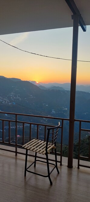 Un banc en bois sur un balcon, offrant une vue sur un coucher de soleil se déclinant sur un paysage montagneux.