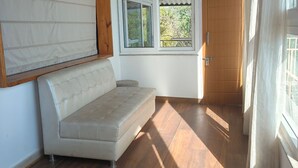 A room with a beige sofa, a wooden ceiling, a window with a view of greenery, and a door. - Saiva Hill Resort (Dehradun)