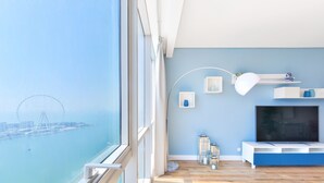 A modern living room with a large window, wooden flooring, a TV, and a blue accent wall. - Luxury JBR - Al Fattan Full Sea View - Free Beach Resorts Access (Dubai)
