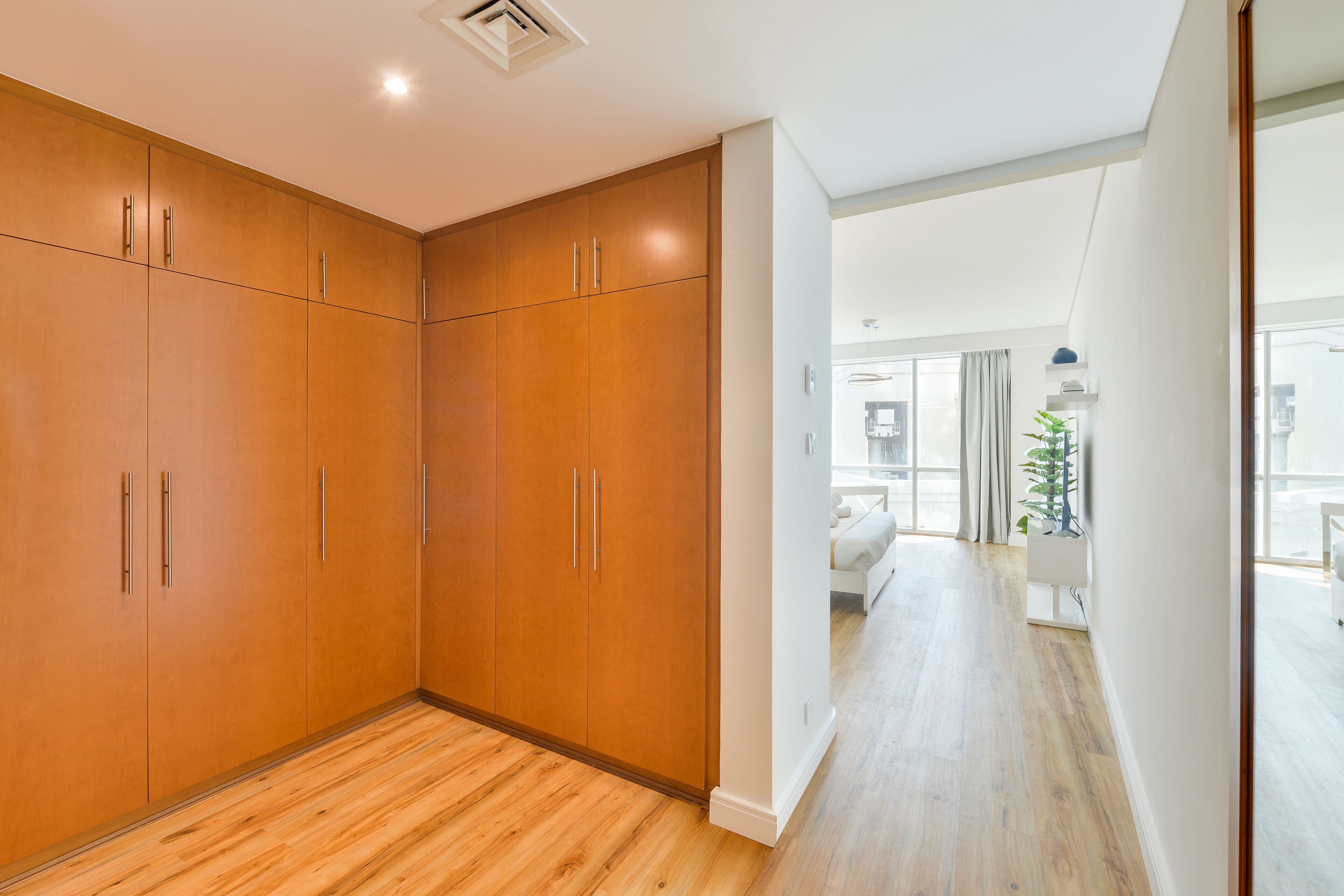 A spacious room with wooden flooring, a large wardrobe with multiple doors, and a view of a living area with a bed and curtains.
