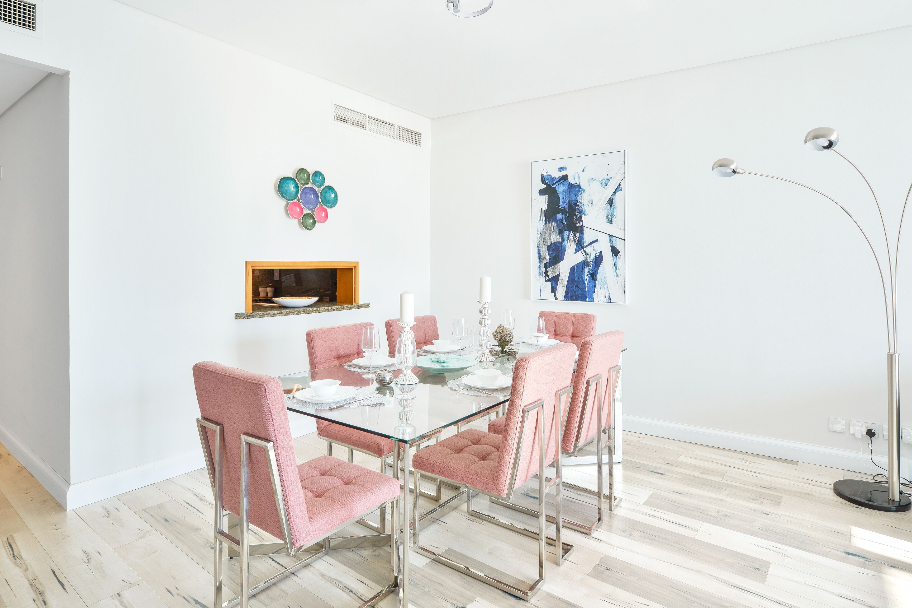 A dining area with a glass table and pink upholstered chairs, a small shelf with decorative items, and a modern painting on the wall.