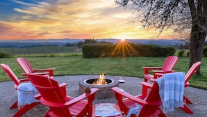 A fire pit with red Adirondack chairs around it, set on a stone patio with a view of the sunset over the hills. - Picturesque Panoramic Vineyard Views From Home and Hot Tub, Pet Friendly, Private, Fire-Pit, BBQ (Newberg)