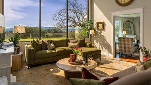 A spacious living room with a large window, a round coffee table, and a comfortable seating area. - Picturesque Panoramic Vineyard Views From Home and Hot Tub, Pet Friendly, Private, Fire-Pit, BBQ (Newberg)