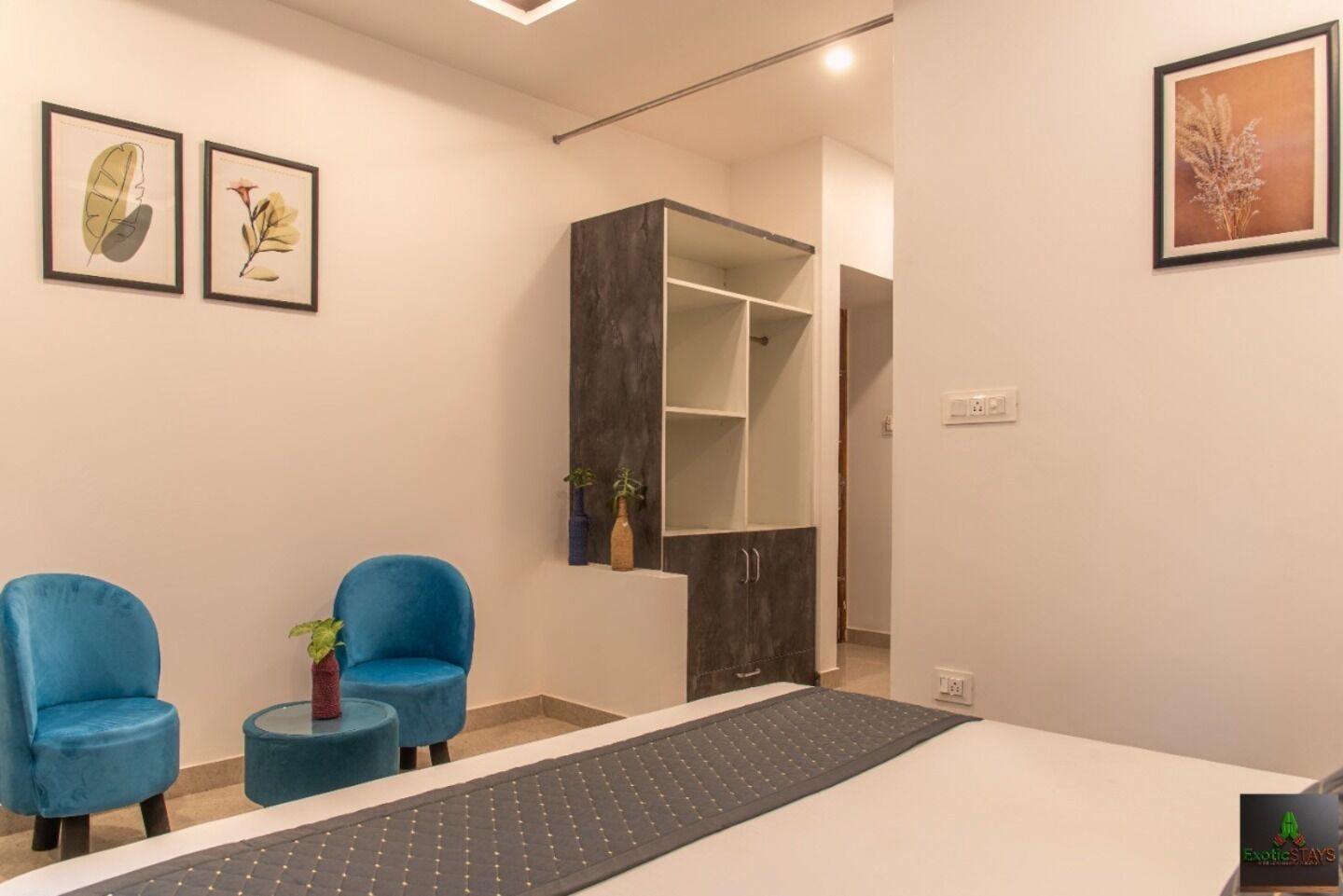 Quarto moderno com escrivaninha, duas cadeiras azuis, uma mesa pequena com uma planta e gravuras botânicas emolduradas na parede.