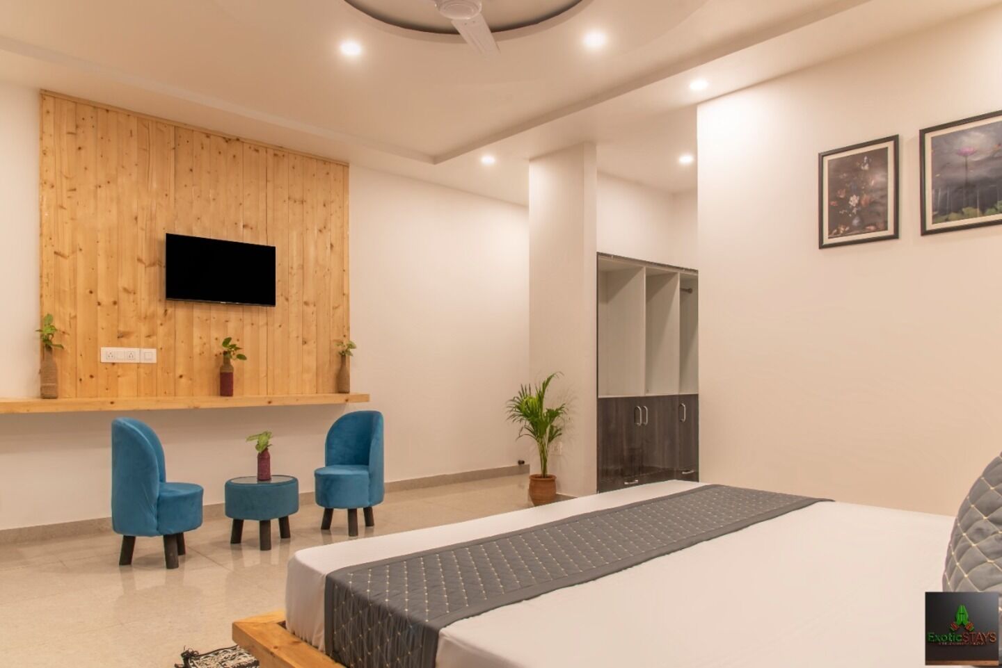 Une chambre d’hôtel moderne avec un grand lit, un téléviseur fixé à un mur en bois, une armoire et deux chaises bleues.