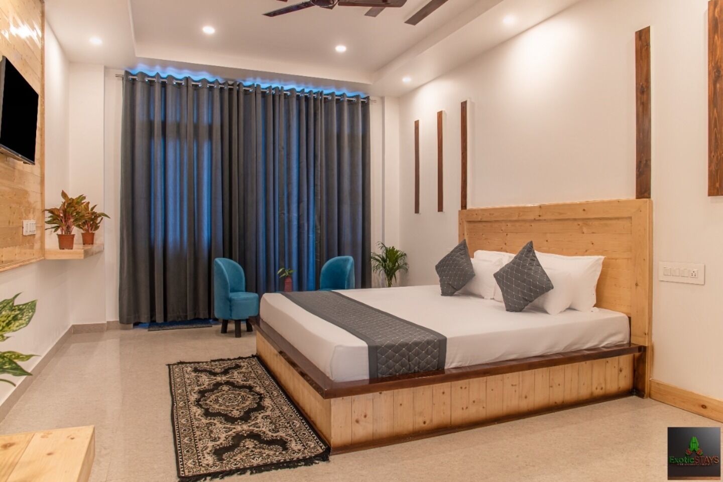 Un dormitorio moderno con cabecera de madera, una cama grande, dos sillas azules, un televisor montado en la pared y una alfombra estampada.