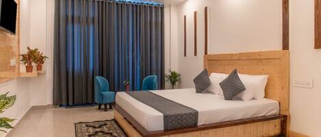 Un dormitorio moderno con cabecera de madera, una cama grande, dos sillas azules, un televisor montado en la pared y una alfombra estampada.