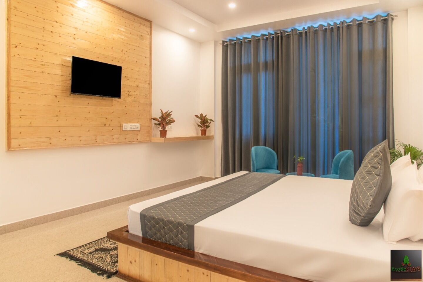 Une chambre d’hôtel moderne avec un mur d’accent en bois, un téléviseur à écran plat, un lit recouvert d’une couette grise, deux chaises et une grande fenêtre avec des rideaux.