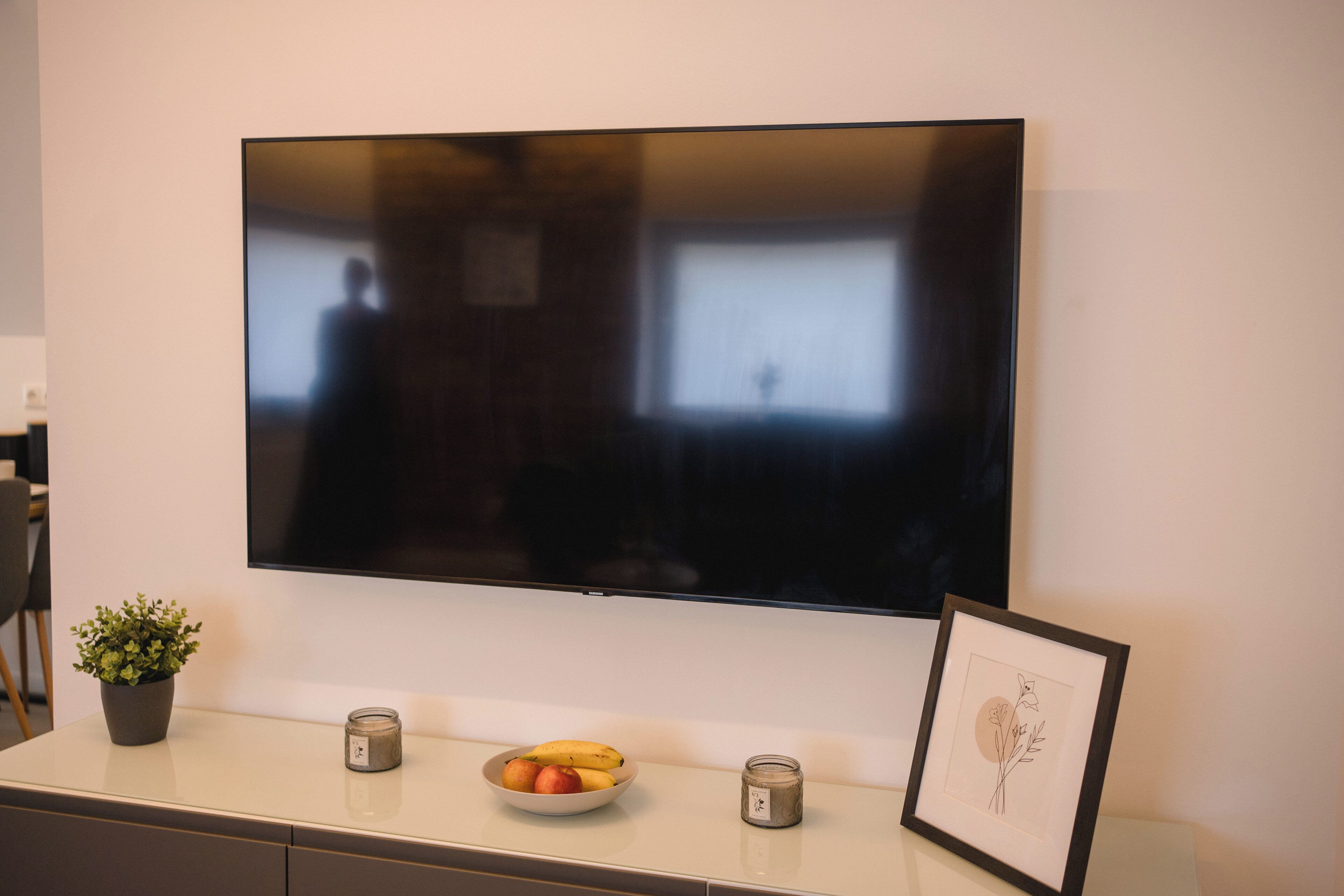 Un téléviseur à écran plat fixé au mur, au-dessus d’une console blanche ornée d’une plante en pot, d’un saladier de fruits et d’un tableau encadré.