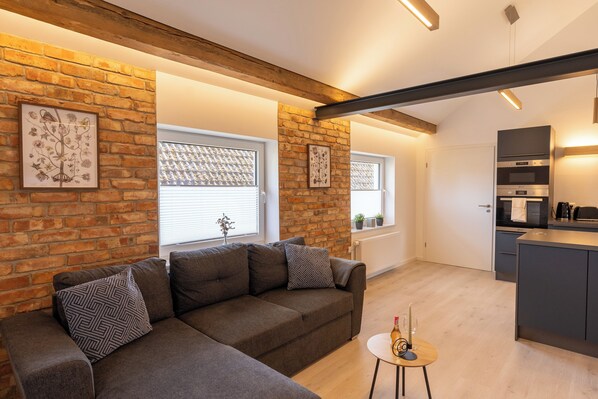 A modern living room with a sectional sofa, a small round table, and a kitchen area with built-in appliances. - Old becomes new (Celle)