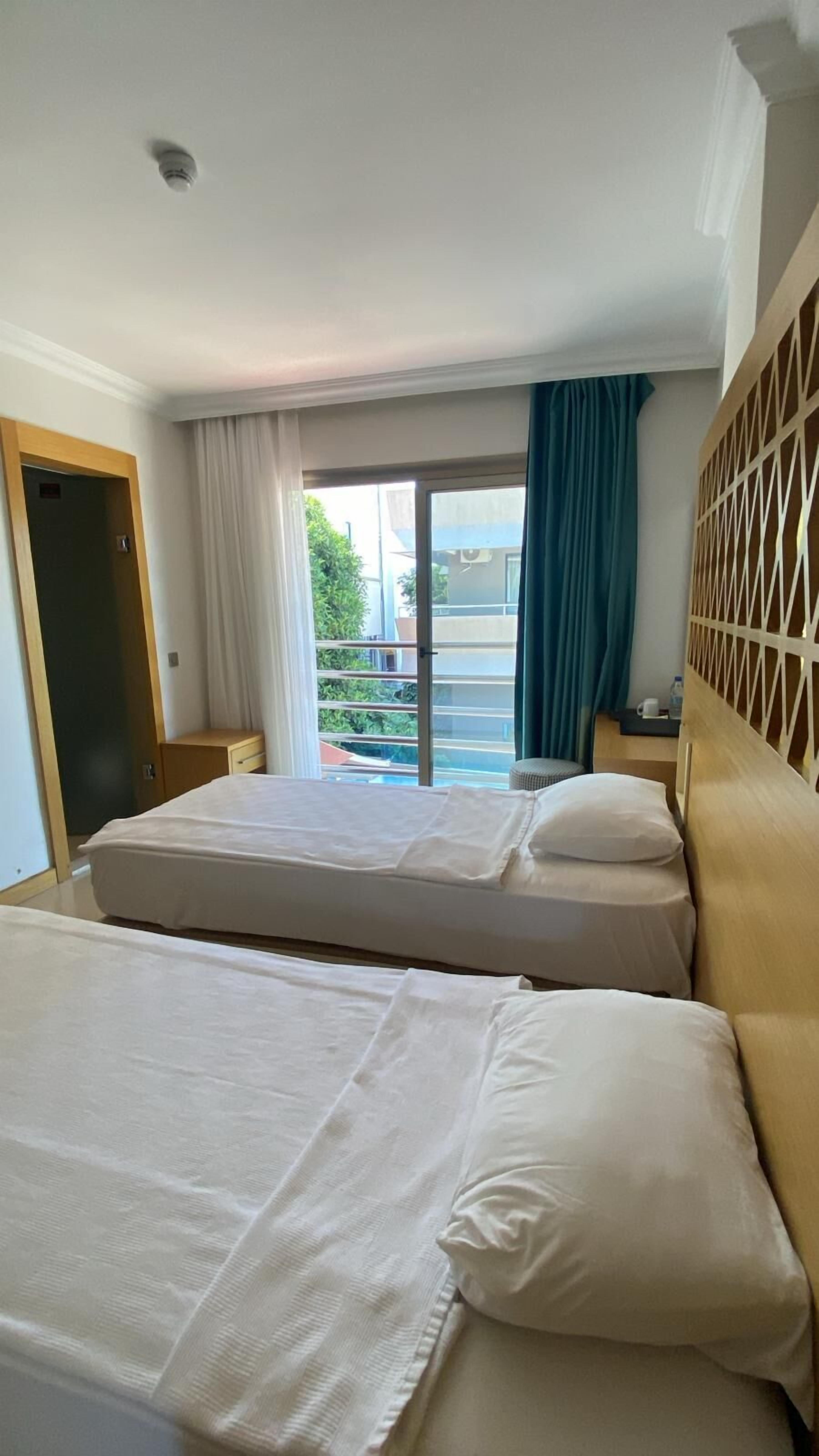 Ein Hotelzimmer mit zwei Betten, Blick ins Freie, einer Tür und einem mit Vorhängen versehenen Fenster.