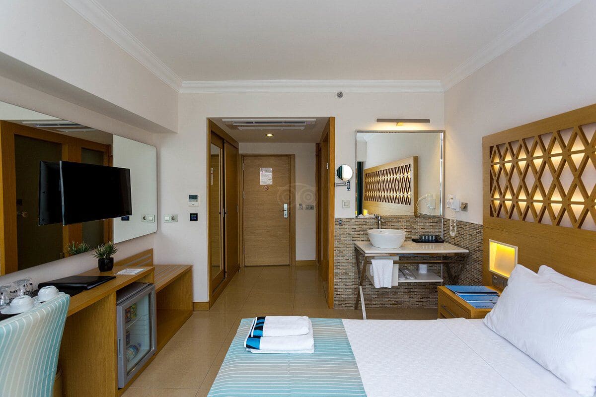 A hotel room with a bed, a desk with a computer, a TV, a bathroom with a sink and mirror, and a wooden headboard.