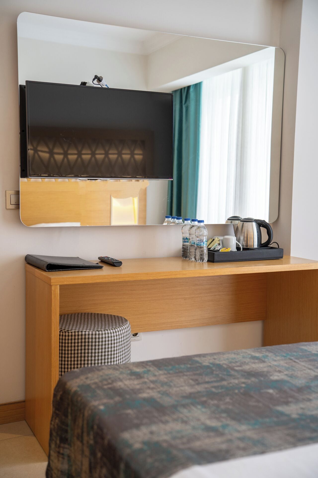 A hotel room with a wooden desk, a bed with a patterned bedspread, a mirror, a kettle, and a tray with bottles and a cup.