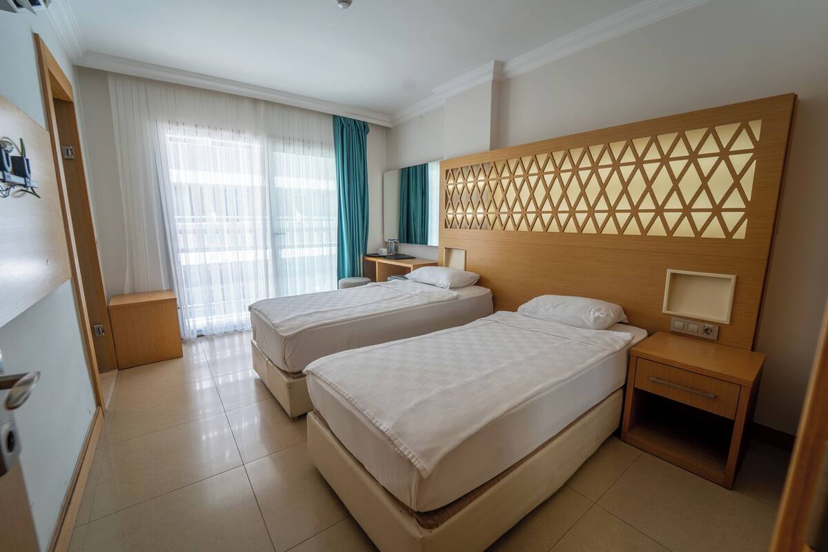 A hotel room with two beds, a wooden headboard, a bedside table with a lamp, and a large window with curtains.