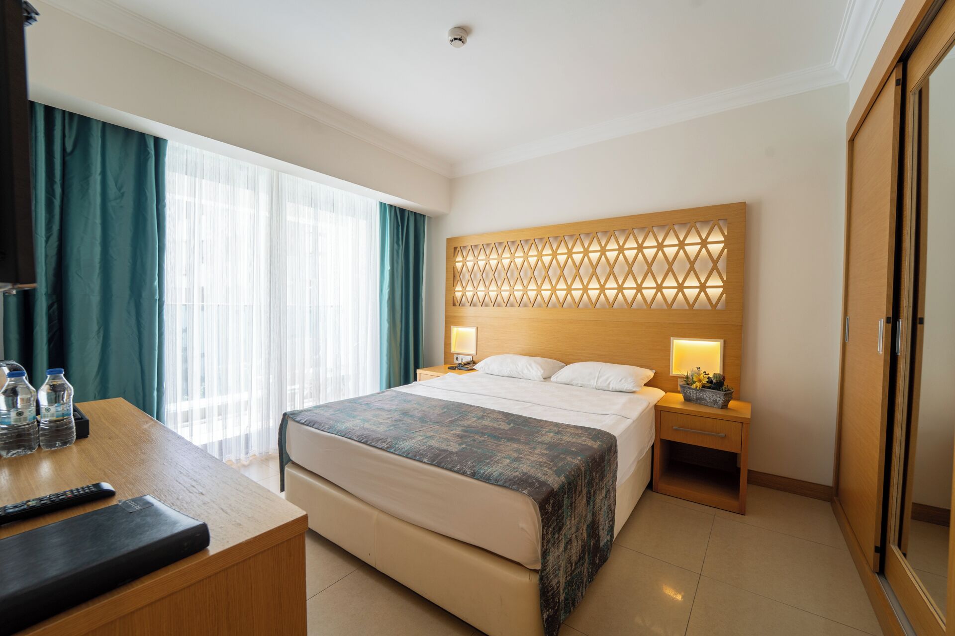 Een hotelkamer met een bed, nachtkastjes, een bureau met stoel, een kledingkast en een raam met gordijnen.