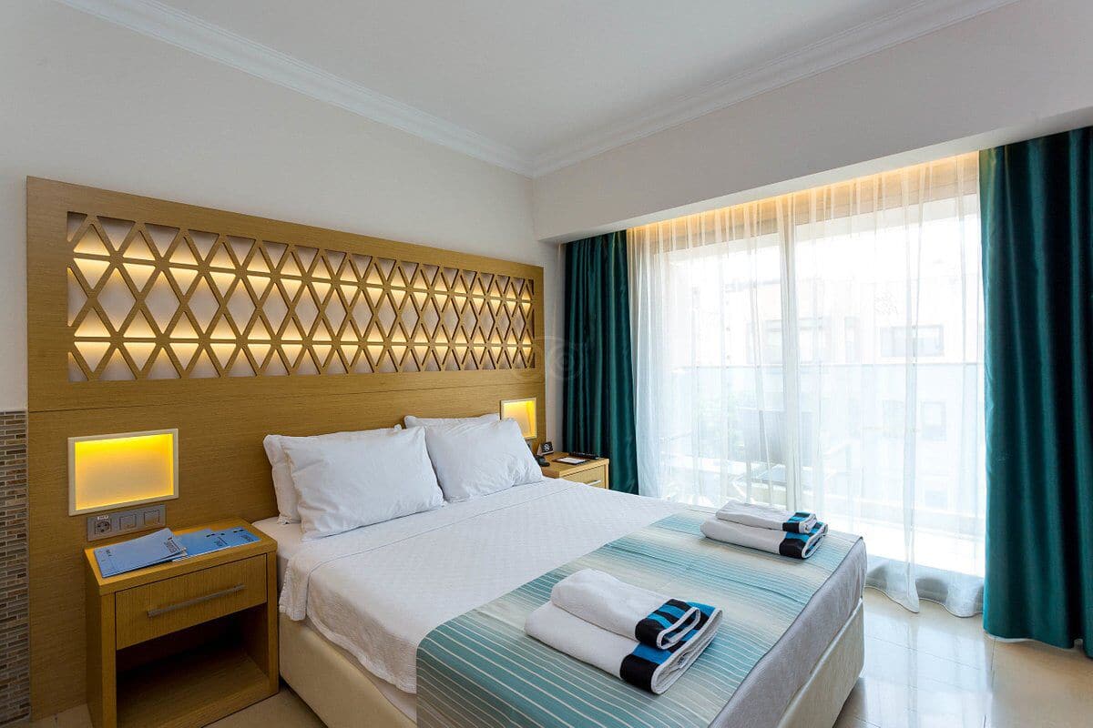 A hotel room with a large bed, a wooden headboard with a geometric pattern, a bedside table with a lamp, and a window with curtains.