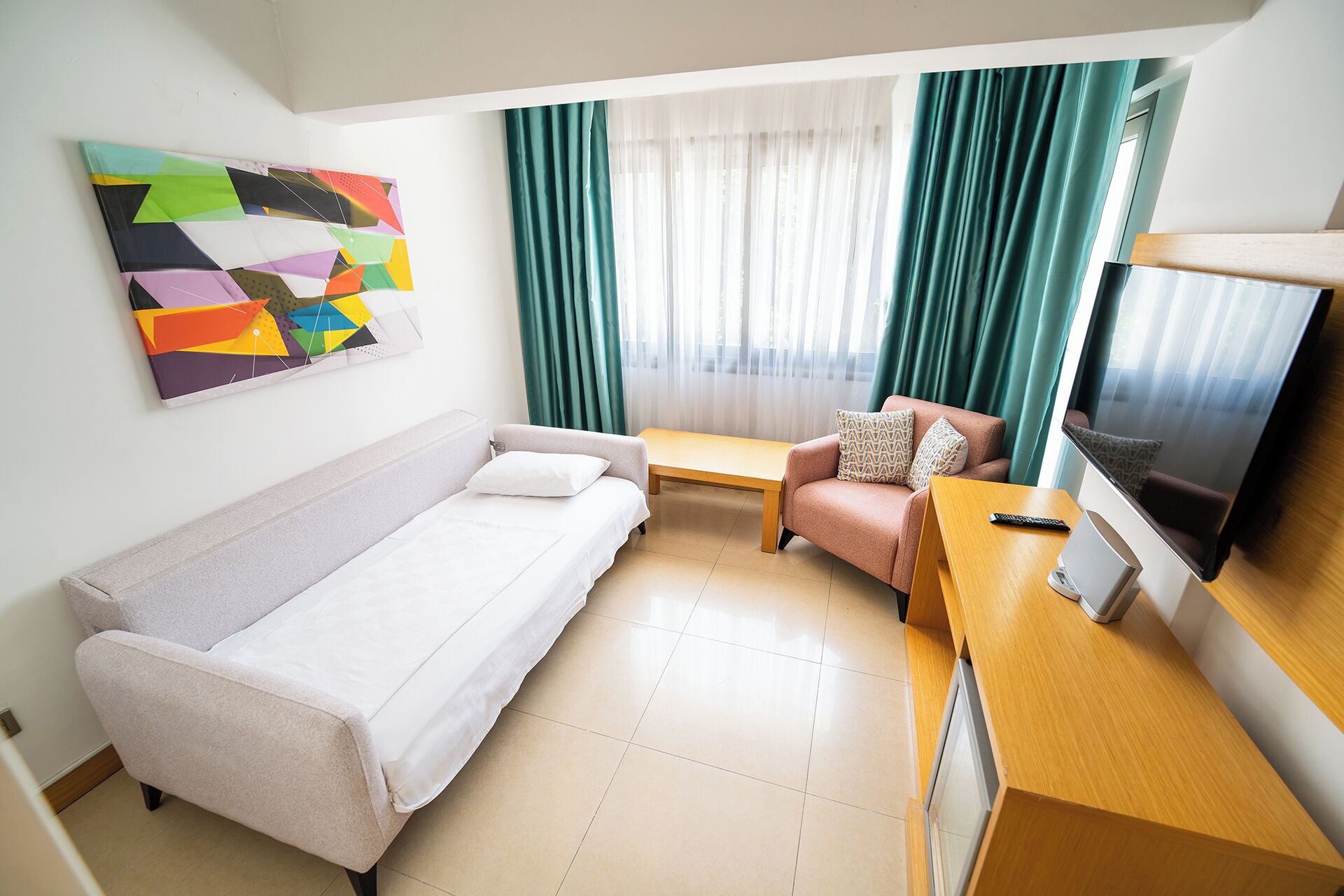 A compact hotel room with a single bed, a sofa, a small table, a TV, and a window with curtains.