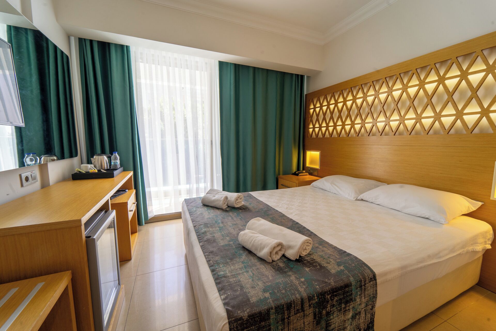 A hotel room with a bed, a desk, a mirror, a nightstand, and a window with curtains.