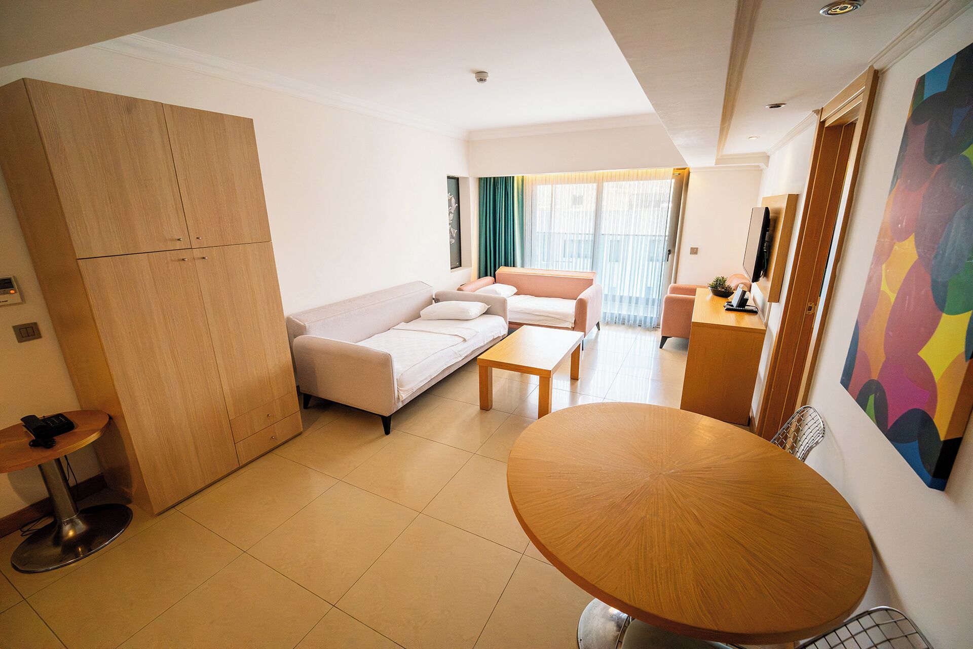 Een moderne hotelkamer met een bank, een eettafel, een kitchenette en een groot raam met gordijnen.