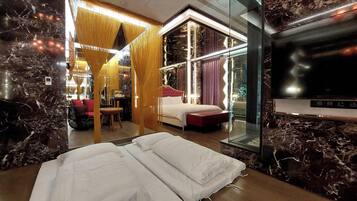 A modern hotel room with a large bed, a sitting area with a red sofa and a small table, and a glass partition separating the bedroom from the living area.