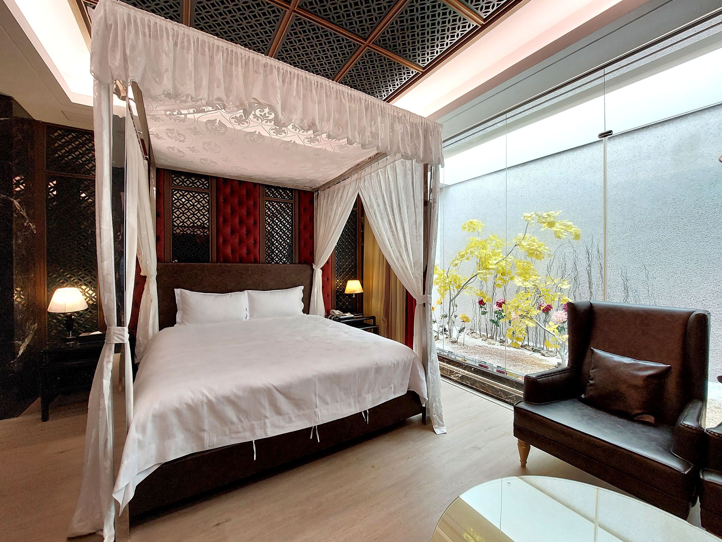 A four-poster bed with a canopy, a sofa, and a view of yellow flowers through a window.
