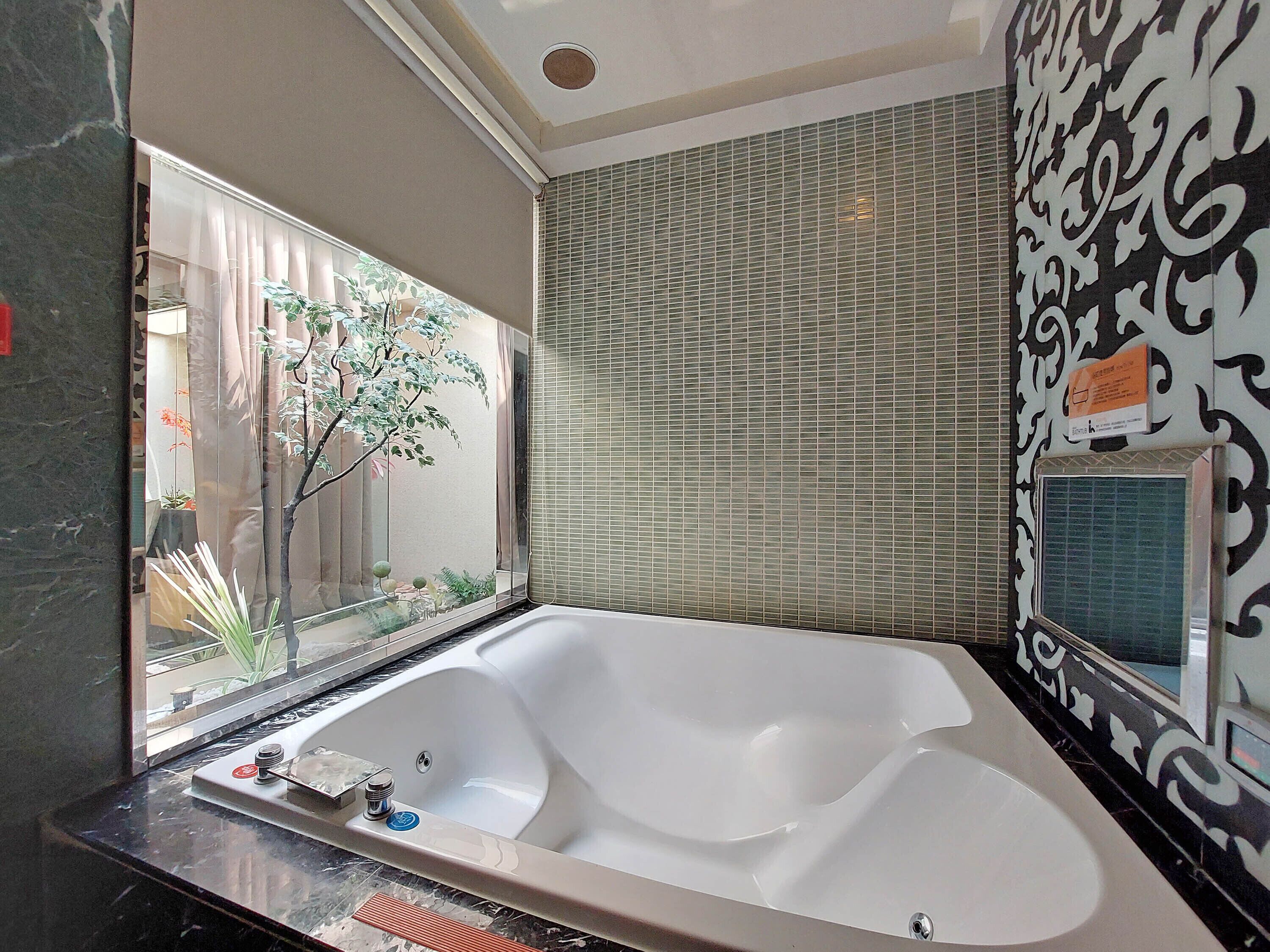 A bathroom with a large bathtub, a window with a view of plants, and a patterned wall.