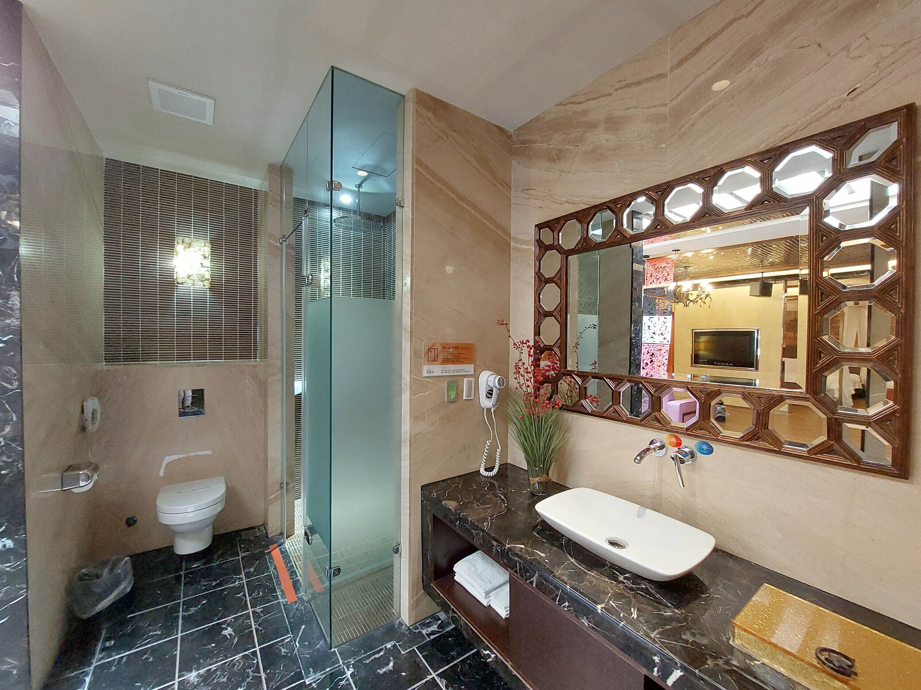 Un baño con cabina de ducha de vidrio, inodoro, lavabo con encimera de mármol y un espejo grande con marco geométrico.