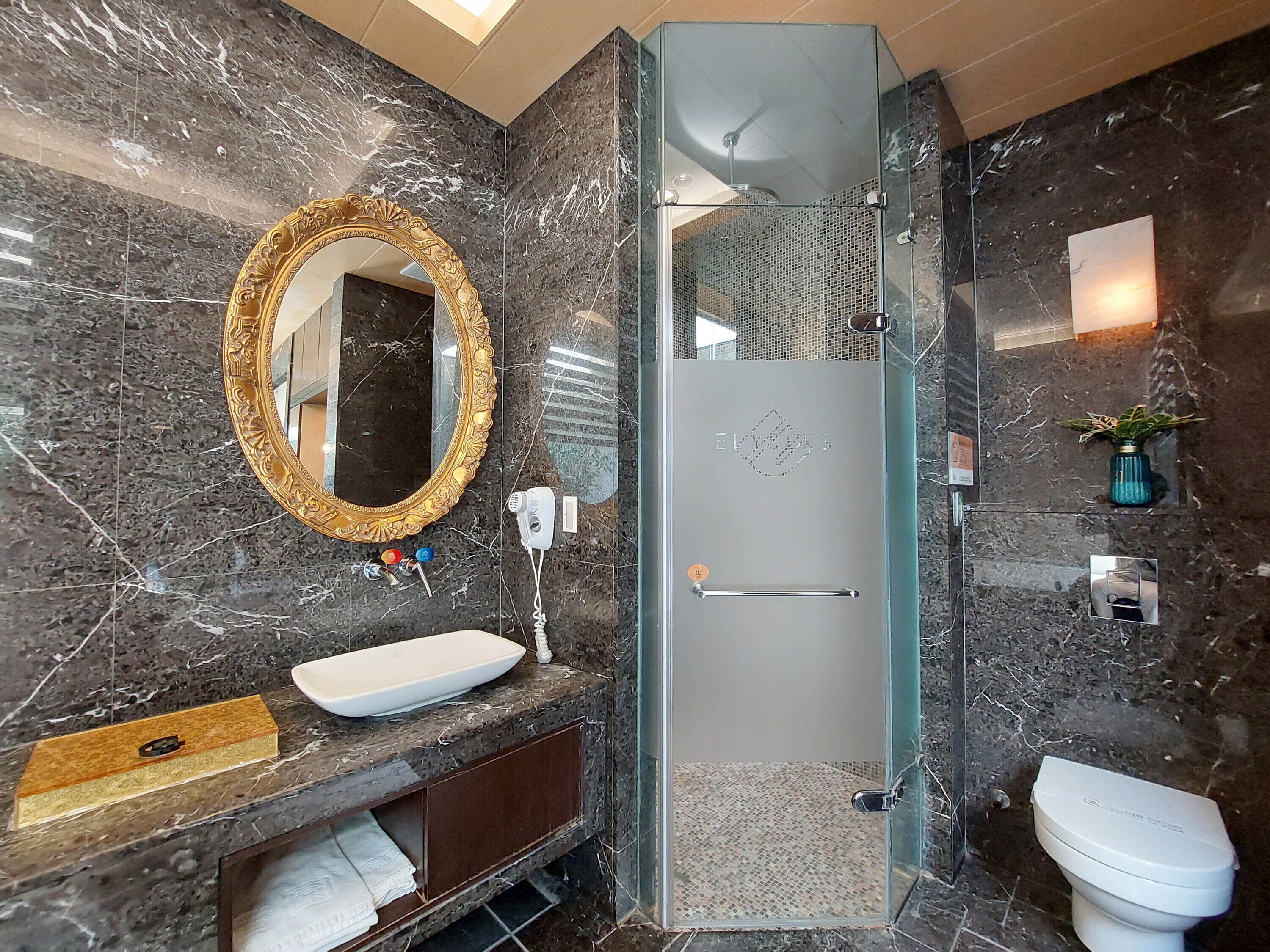A modern bathroom with a marble vanity, a gold-framed mirror, a glass-enclosed shower, and a white toilet.