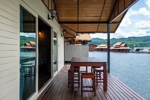 A wooden deck with a table and chairs, overlooking a body of water with boats and thatched-roof buildings in the background.