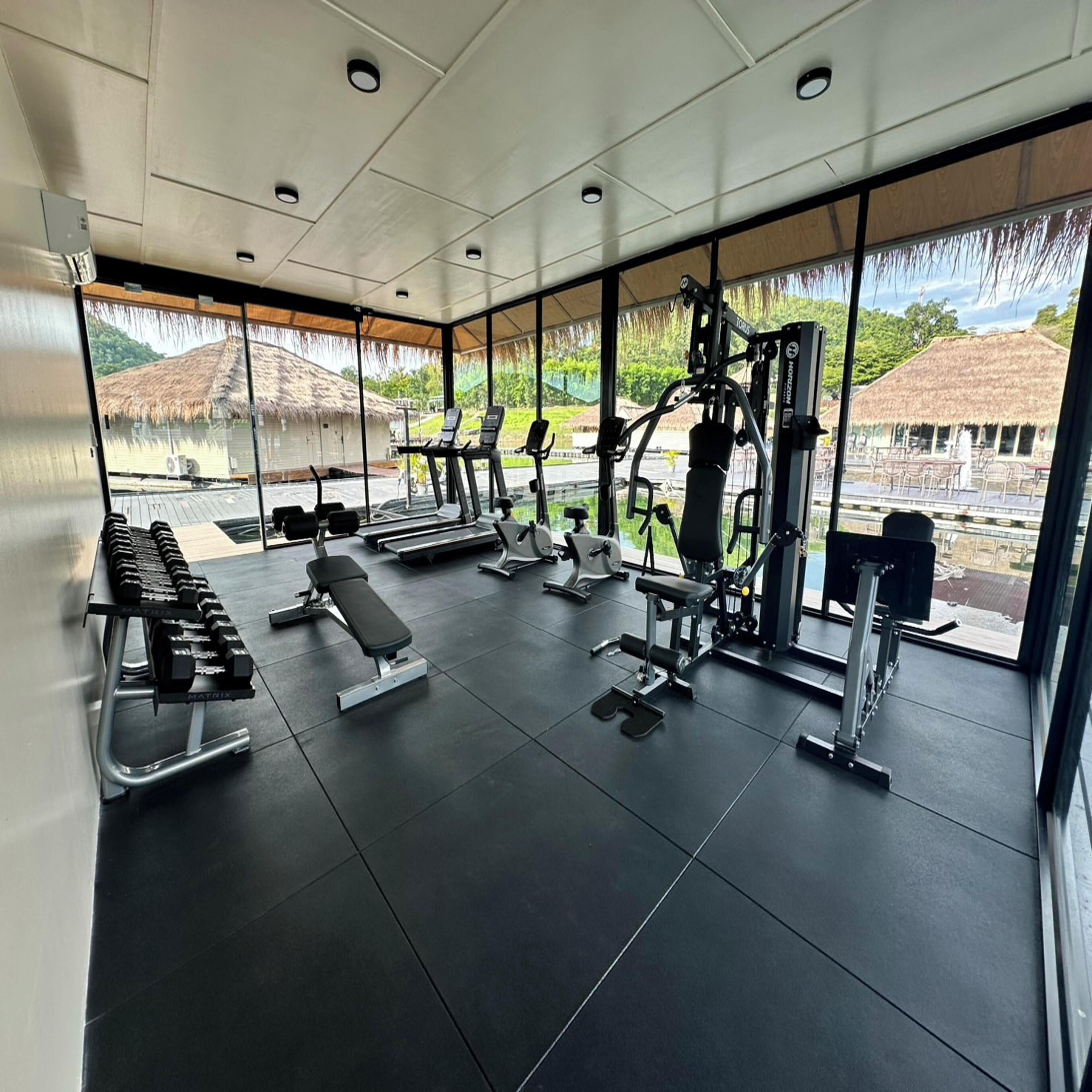 A modern gym with various exercise equipment, large windows, and a view of a thatched-roof building.