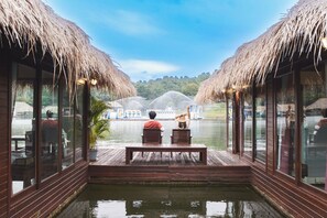 A wooden deck with thatched roofs overlooking a body of water, with a fountain in the background.