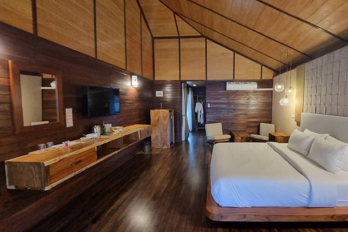 A modern hotel room with a wooden ceiling, a large bed, a TV, a wooden bench, and a mirror.