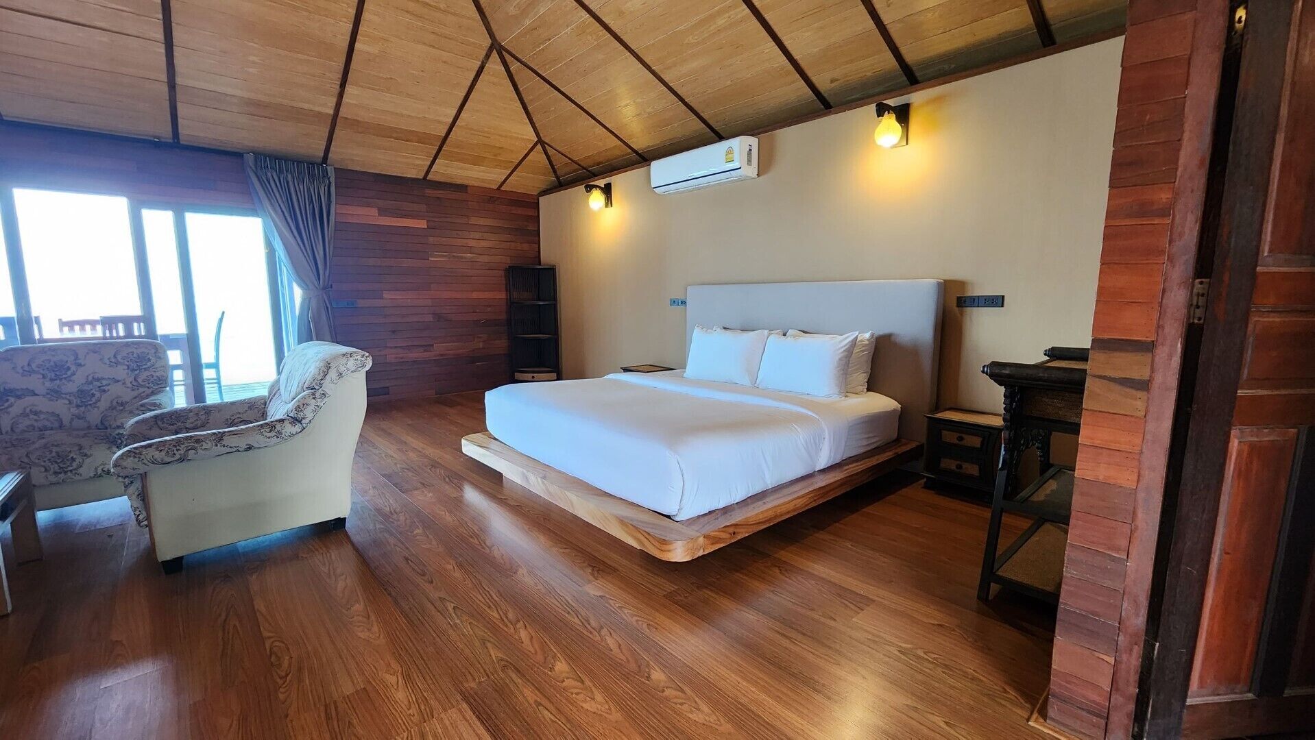 A bedroom with a bed, a chair, wooden flooring, a window with curtains, and a wall-mounted air conditioner.