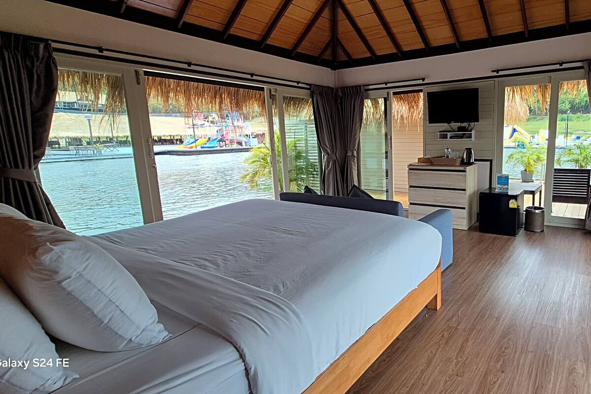 A modern bedroom with a large bed, a desk, and a TV. It has large windows with a view of a water body and a playground.
